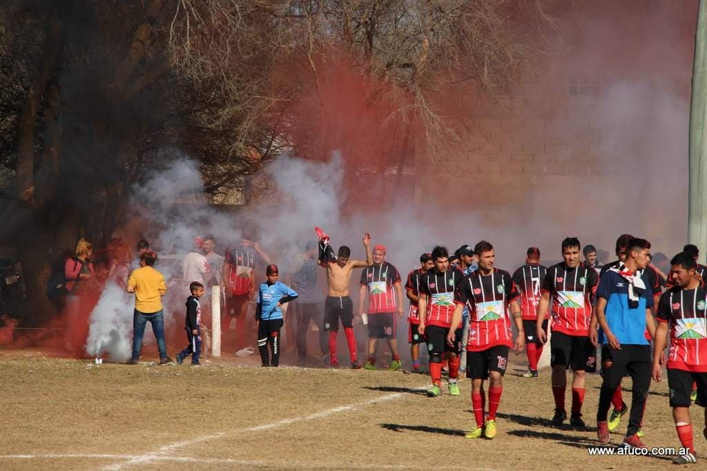 Jugadores de un equipo de fútbol comercial, con camisetas a rayas rojas y negras, salen a la cancha entre humo de colores y el público.