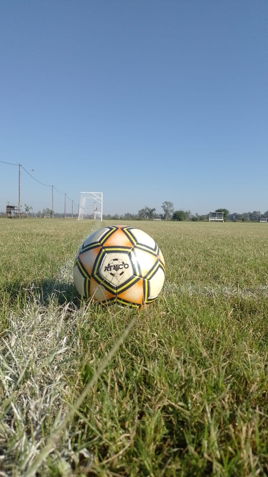 Pelota con el nombre AFUCO sobre la línea de cal de una cancha de fútbol, con un arco al fondo.